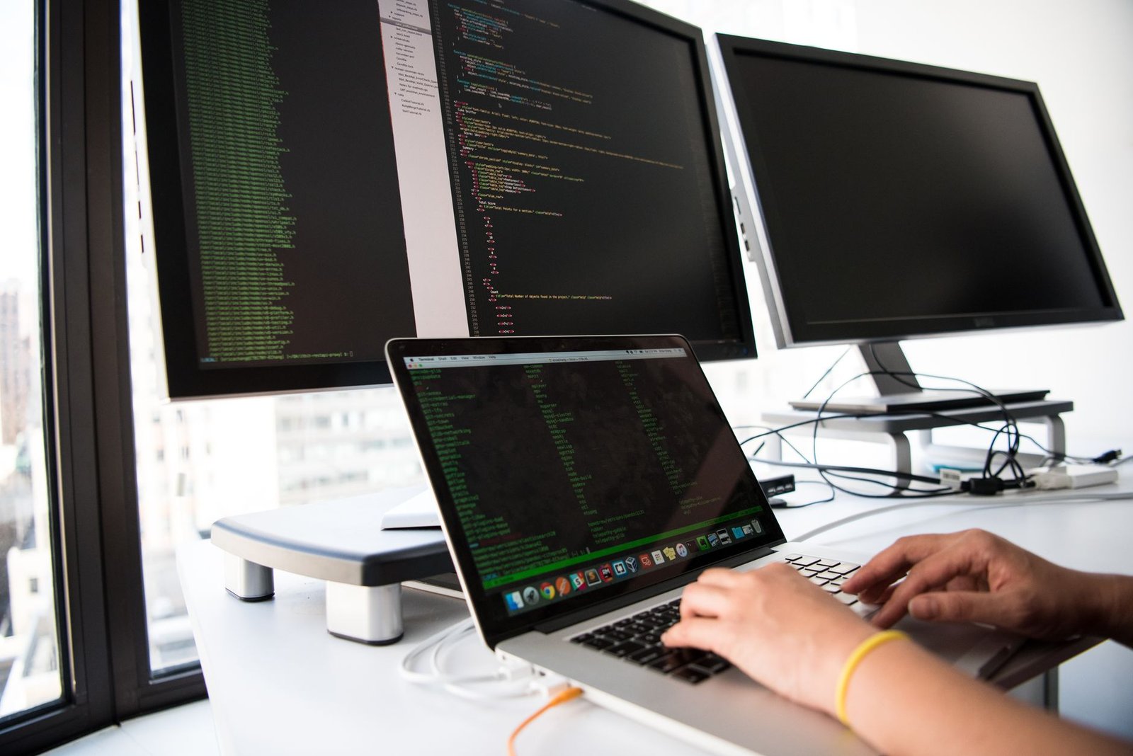 A software engineer actively writing code and reviewing architectural diagrams on a multi-monitor workstation in A modern, well-lit UK tech startup engineering office with exposed brick walls, a standing desk, and subtle background hints of London architecture through a large window